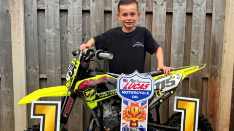 A young boy in a black t-shirt smiles sweetly as he stands behind a yellow motorbike