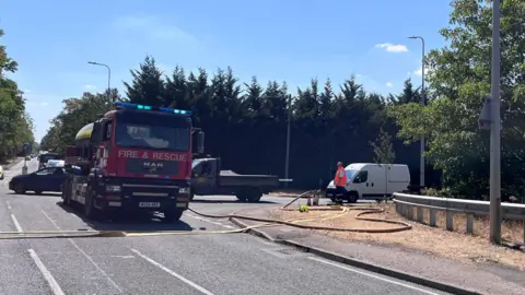 Nicola Haseler/BBC A fire engine on a road with a yellow hose pipe coming out of it. There is a white van to the right, and a man by it. There are trees behind the vehicles and a path to the right. 