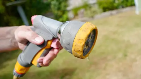 A hand holding the end of a hosepipe which is dripping with water. A garden which is blurred can be seen in the background. 