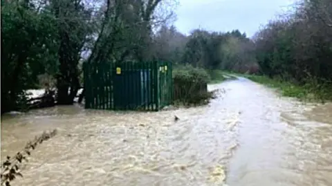 Tim Field A grainy photograph of a flooded country lane. The water looks deep and fast moving in places. There is a small electricity substation which is surrounded by a green metal fence with yellow warning signs attached. It appears the subsataion is flooded at ground level.