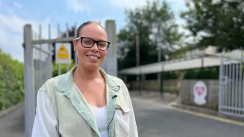 Mother Alicia Bailey stands outside the school gates. She has glasses on and her hair tied back and is wearing a white vest with a green oversized shirt on top.