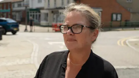 Ant Saddington/BBC A woman with glasses, wearing a black dress stands on Buntingford high street.