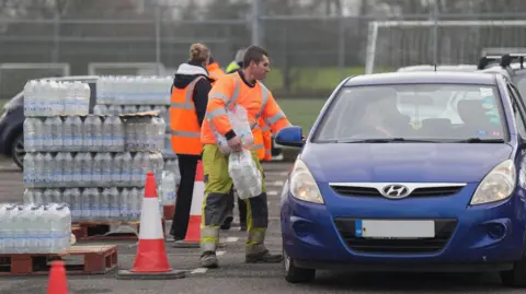A man in his-vis clothing carrying packs of bottles of water in a car park. He is stood beside and looking into a blue car on the right, which is facing towards the camera. A woman is stood behind him, facing away, looking at a pallet stacked high with bottles of water.