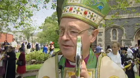Former Bishop of Swansea and Brecon Anthony Pierce is a grey-haired man wearing thin-rimmed rectangular glasses. He is wearing a purple shirt with a dog collar and has green, white and gold robes. 