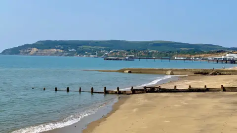 Wight Life A empty sandy beach and sea on a sunny day with cliffs and green hills in the background.