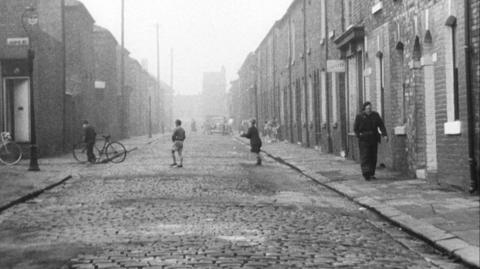 A black and white wide shot of a street of terraced houses. Children are playing on the road and a man is walking on the footpath.