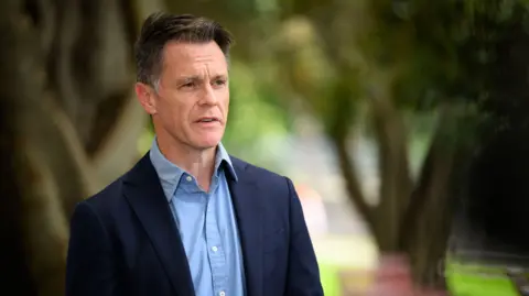 Chris Minns speaks at a press conference, wearing a navy jacket and blue collared shirt. He has brown hair with grey tips.