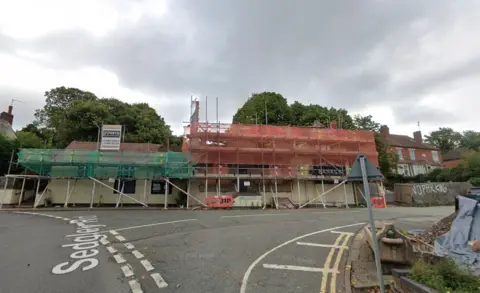 A long building which has scaffolding and netting over it. It is on the corner of a road. 