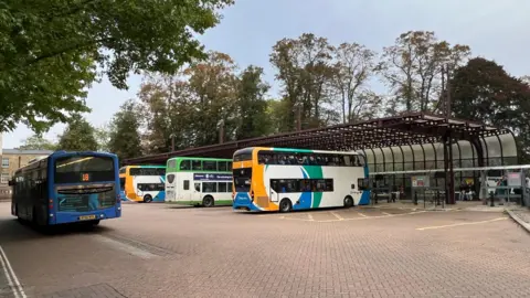 Three parked double-decker buses at the central bus depot in Cambridge. A fourth single-decker bus is to the left. A number of tall trees are in the background.