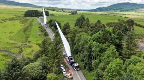 An aerial view of the A76 through southern Scotland with two large turbine carriers travelling in convoy. The road is surrounded by trees and hills and there are workers in orange outfits standing on the road in front of the lead vehicle. Cars are tailed back behind the second vehicle in the convoy.