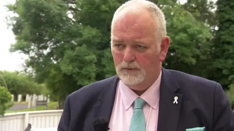 Jonathan Evison with a goatee-style beard standing in front of trees wearing a dark jacket and pink shirt. 