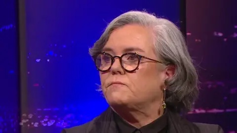 RTE Rosie O'Donnell looks to her right while on the Late, Late Show. She has grey hair down to her shoulders and black-rimmed glasses and is wearing a dark jacket and shirt