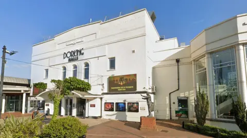 Google The outside of a theatre. The building is white with film posters on the outside and black letters that spell 'Dorking Halls'. 