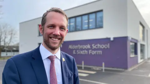 A man with light coloured hair, a beard and blue eyes smiles into the camera. He is wearing a smart blue jacket, light blue shirt and a patterned tie. Behind is a purple and white building with Alderbrook School & Sixth Form written on it.