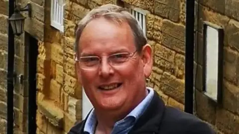 Simon Jones Simon Jones is wearing a dark navy coat over a blue shirt.