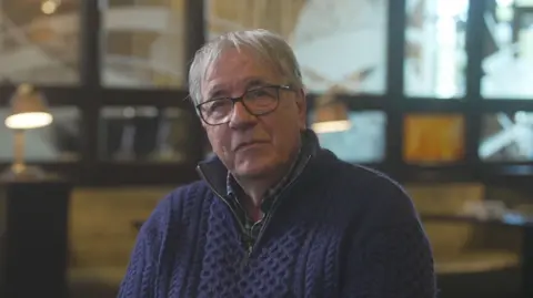 A man with grey hair is sat looking at the camera. He is wearing glasses and a thick, navy twead jumper with a quarter zip. Behind the man is an out of focus industrial space with lots of windows and open space. There are some lamps placed in front of the windows. 