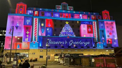 An image of the projected film onto the side of the large Queen's Hotel in Leeds. The image shows an advent calendar with a Christmas tree in the middle, in pink, purple, red and blue. 