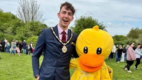 supplied Harry stands in a navy suit and pink shirt with a burgundy tie and a gold chain around his neck. He is laughing and has his arm around someone in a bright yellow duck outfit.