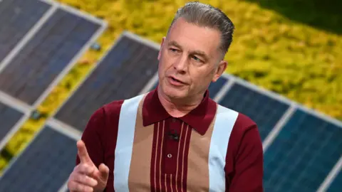 BBC Chris Packham sits in a television studio. He is wearing a burgundy T-shirt with a collar, with blue and beige stripes down the front.