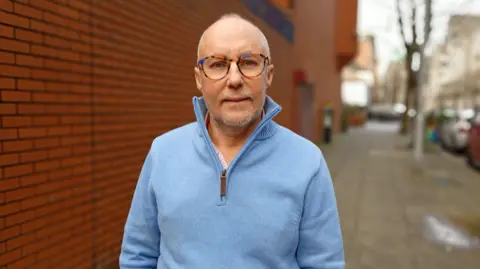 John Miskelly is wearing a pale blue quarter zip jumper and multicoloured rimmed glasses. He is staring at the camera while standing on a street with a bed brick wall next to him. The background is out of focus.