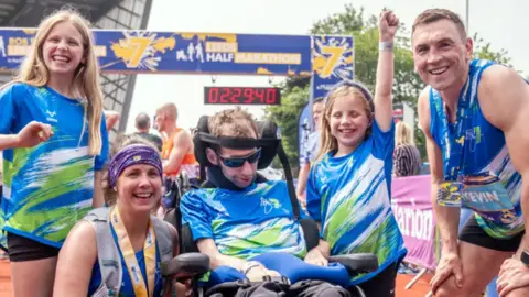 PA Media Rob Burrow in a wheelchair surrounded by his smiling wife, his two daughters and Sinfield at the Leeds Marathon