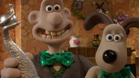 PA Image of clay modelled characters Wallace and his dog Gromit. Wallace is holding a silver trophy. They are both wearing green shiny green bow ties. In the background can be seen a tile fireplace, oval mirror above the fire and a china and glass vase.