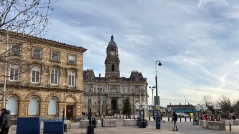 Gemma Dillon/BBC Image shows Dewsbury town centre. 
The town hall is in the background, a big stone building.
To the left of shot is also a boarded up , stone building which used to be Barclays bank but has stood empty for the last two years. 