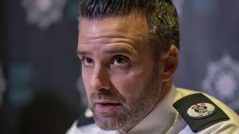 PA A police officer in close up wearing his uniform shirt, with epaulettes on the shoulders. He has short, thick salt and pepper hair and stubble. 