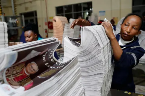 THOMAS MUKOYA/REUTERS A woman next to a stack of newspapers with Pope Leo XIV on the front page. 
