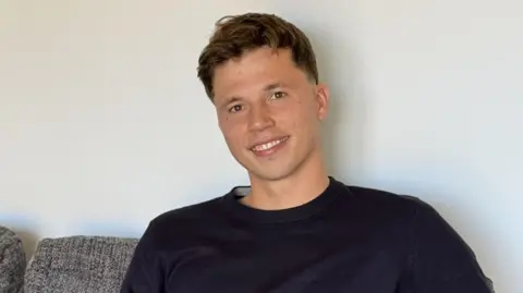 Freddie Woodward: a young man with short dark hair sits on a grey sofa, smiling at the camera. He has short brown hair and wears a navy jumper.