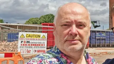 Ian Hughes A man with short grey hair and a floral blue shirt standing in front of a building site