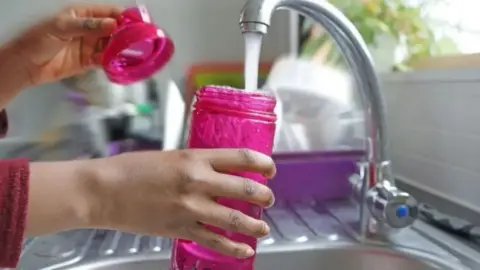 Silver metal sink with tap and woman's hand holding a pink water bottle under the running water.