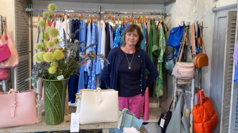 Corinne Miles standing in front of a row of clothes in her shop, behind a table holding handbags and flowers