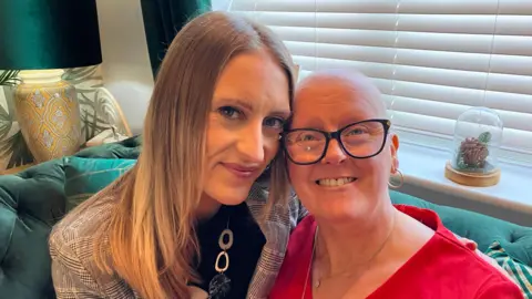 Mo Jackson, smiling and wearing a red top, sits with her 36-year-old daughter Amy on a sofa.