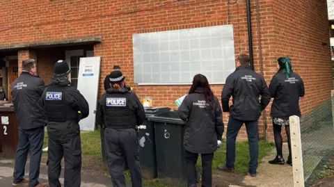 Melton Borough Council Police officers and council staff stand in front of a house with a silver steel place secured over the window.