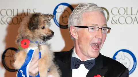 A silver haired Paul O'Grady wearing glasses and a black jacket with bow tie, carrying a dog with a blue collar and both are wearing red poppies. They are standing infront of a sign at a press-call 