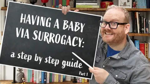 Kevin Pittuck-Bennett A smiling Kevin holds up a blackboard that reads: "Having a baby via surrogacy: a step by step guide". In the background is a bookshelf with lots of different coloured books. 