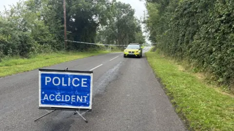 BBC A rural road with a blue sign reading "police accident". There is a PSNI car parked on the road in the distance. 