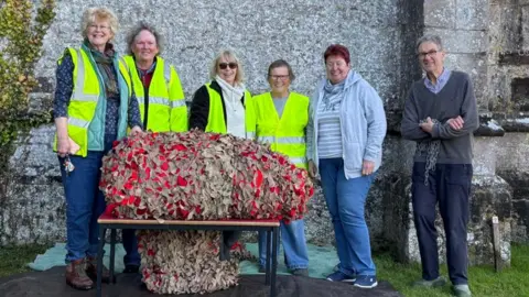 Guerrilla Gardeners Six people standing outside the church in Wool next to a table with the folded up cargo net on top, bulging with red poppies.