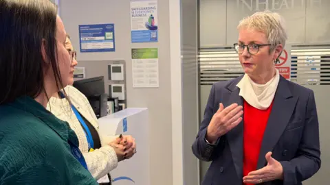 Karin Smyth on the right speaking to two women on the left. She has short hair and is wearing a red top with a dark blue blazer. She is also wearing glasses. 