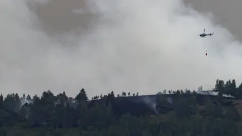 Reuters A helicopter in mid-air against a background of a plume of white smoke over a dark landscape.