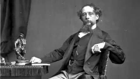 Getty Images Charles Dickens sat at a desk. He has a long beard, is wearing a suit, and there is a metal statue and a book on the table in the black and white image.