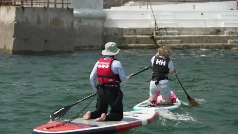 Two women kneeling on paddleboards paddle away from the camera. They are both wearing buoyancy aids and have waist leashes attaching them to their boards.