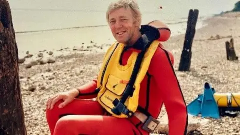 A man with fair hair sitting on a pebble beach. He has a red wetsuit on and a yellow life vest over the top. He is smiling as he looks into the camera, sideways facing.