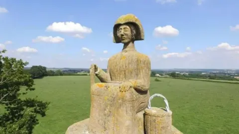 A stone statue of a woman with a shopping basket wearing a bonnet, with fields behind and tree.