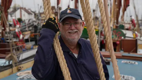 John Fairhall/BBC Tim Kenney in navy blue overalls, yellow and black gloves, and a navy baseball cap, leaning on the ropes aboard Pudge the sailing barge.
