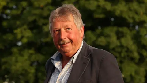 PA Media Sammy Wilson has dark grey hair and a moustache and is wearing a black suit jacket and open-necked shirt