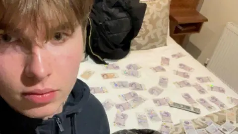 Counter Terrorism Policing A boy with his face near the camera and a hotel bed in the background with lots of £20 notes spread out on it. 