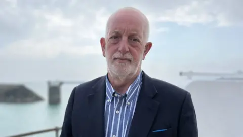 Steve Luce pictured with the sea behind him. He's wearing a blue striped shirt and navy jacket. He has short white hair in a buzz cut and short white stubble. 