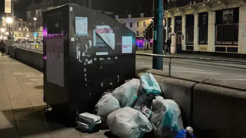 Kieran Hunt White bin bags piled beside a large black public waste unit on a lit Margate pavement at night, with rubbish stacked against a low wall